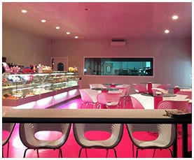 La cafetería de Ajalvir: barra iluminada en fucsia, taburetes y mesas junto a la vitrina
