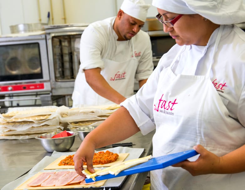 Dos pasteleros montando empanadas de hojaldre a mano sobre la mesa del obrador, con el logotipo «fast pan & pasteles» bordado en el delantal